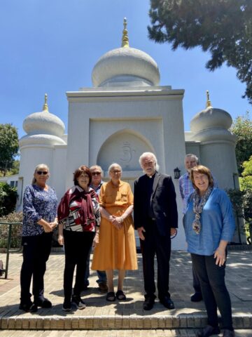 Group of eight adults posing in front of a white temple with onion-shaped domes and gold spires under a clear blue sky.