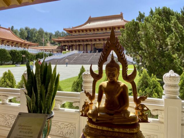 Bronze Buddha statue with ornate flame halo in a temple courtyard, orange-roofed buildings and green trees in the background under a blue sky.