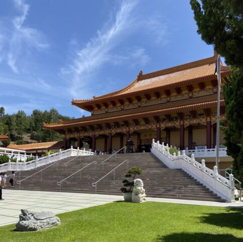 Ornate Asian temple facade with layered orange-tiled roof, red columns, and a stone dragon statue on the lawn under a clear blue sky.