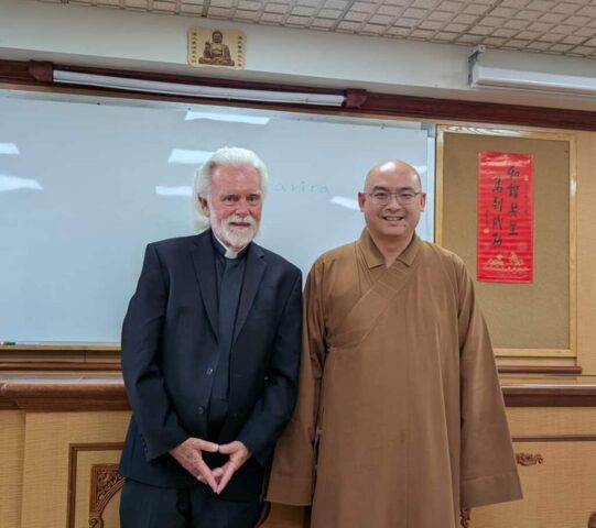 Two men posing for a photo in a classroom: one in a black suit with a clerical collar and the other in brown Buddhist robes, standing in front of a whiteboard and a red Chinese scroll on the wall behind them.