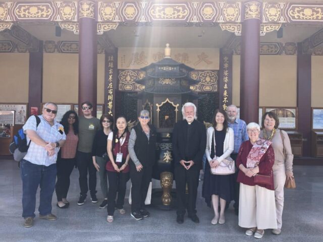 Group of diverse adults posing in front of an ornate Asian temple entrance with gold detailing and carved pillars.