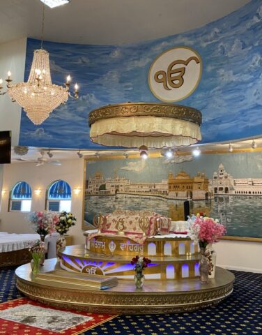 Ornate circular altar with gold trim and canopy inside a temple hall, with a crystal chandelier and a blue mural backdrop.