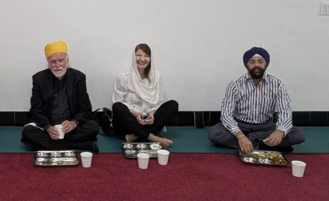 Three adults sit cross-legged on a red carpet, with metal snack trays and paper cups in front; left man in a yellow turban, center woman in white, right man in blue turban.