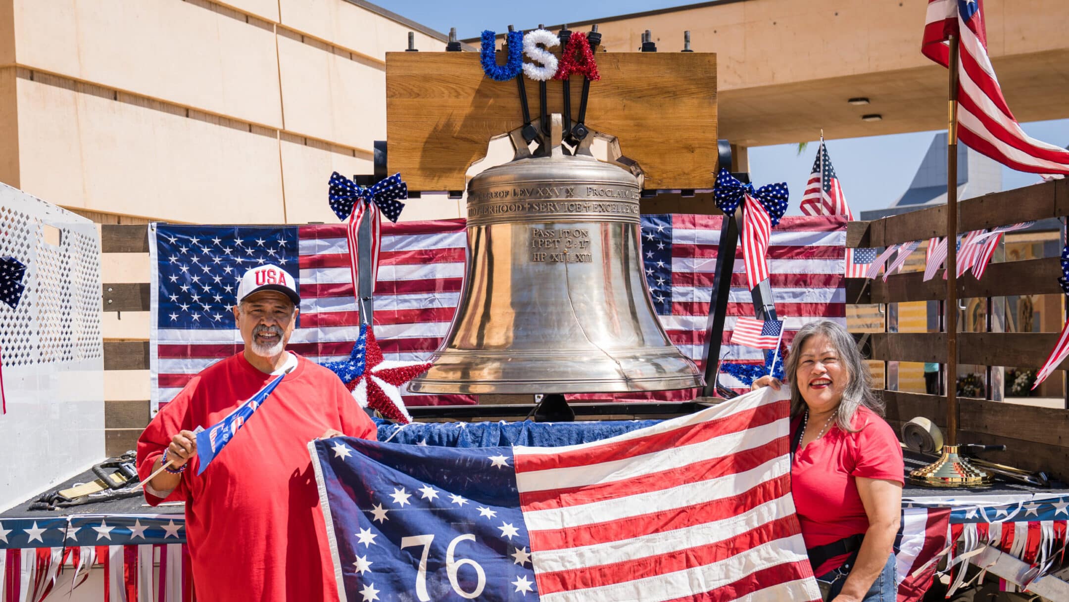Man and woman in red shirts pose beside a large bell on a patriotic parade float with American flags and bunting.