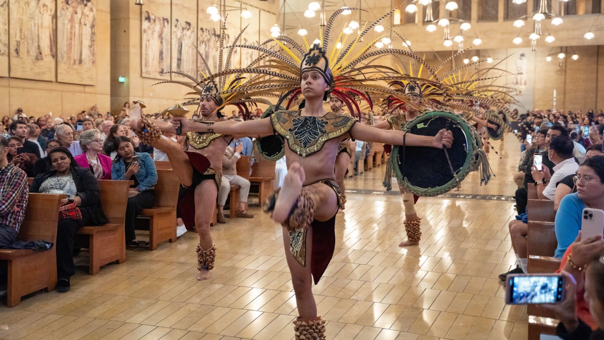 Dancers in ornate costumes with large feather headdresses perform on a church-like hall floor as a seated audience watches and records the moment.