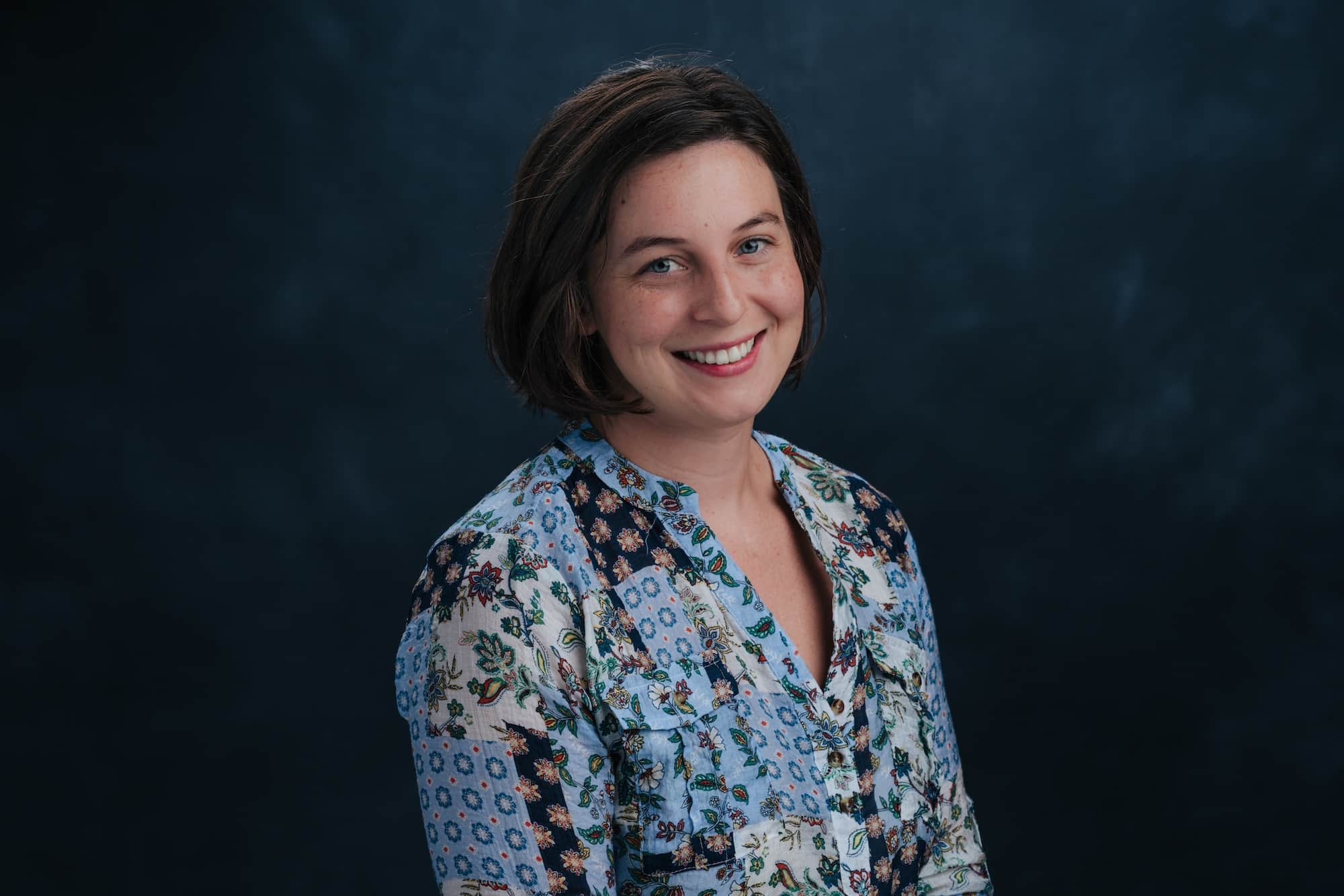 Smiling woman with short brown hair in a light blue floral blouse against a dark blue backdrop.