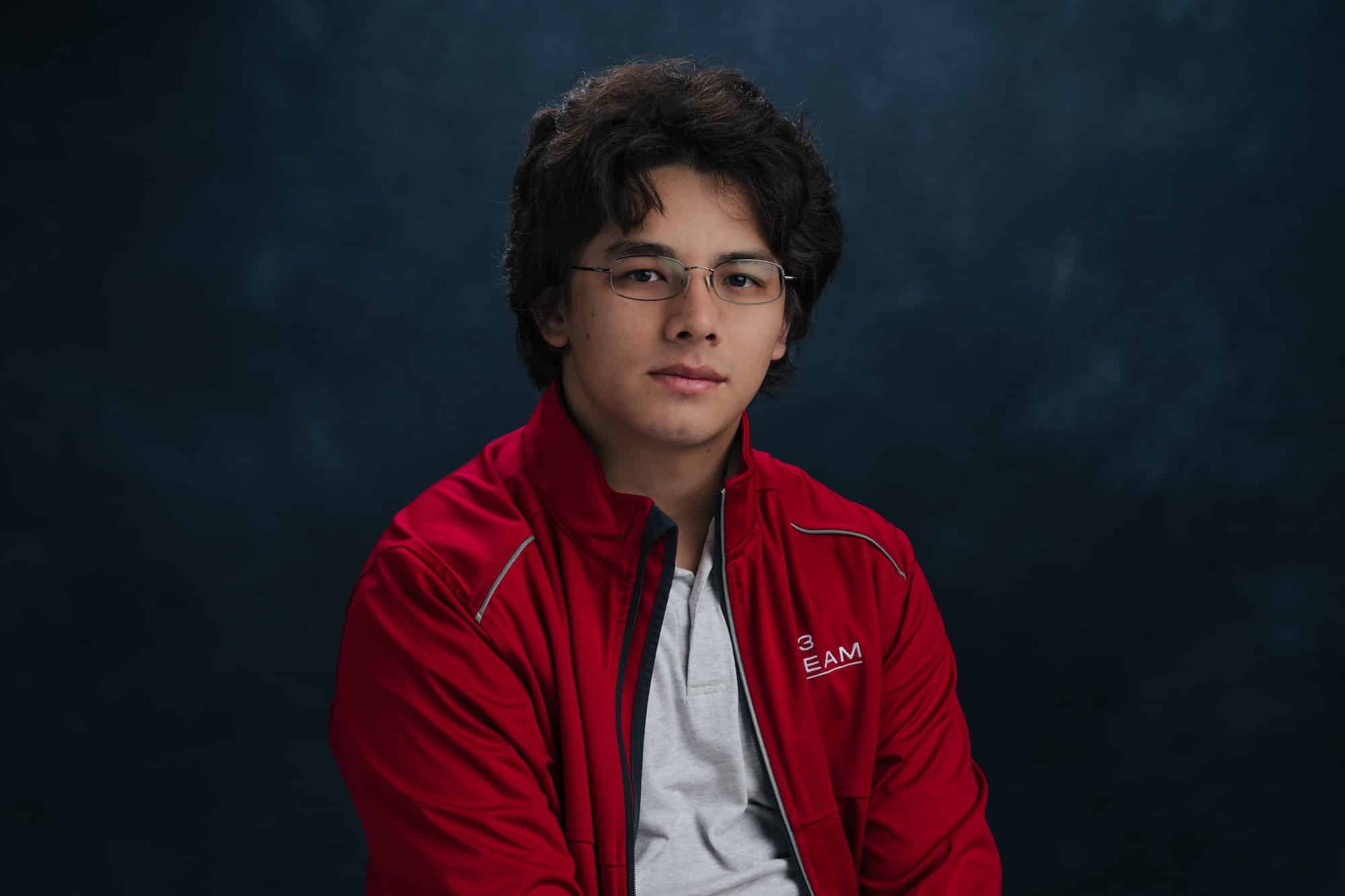 Portrait of a young man with dark hair and glasses, wearing a red jacket over a gray shirt, against a dark blue backdrop.