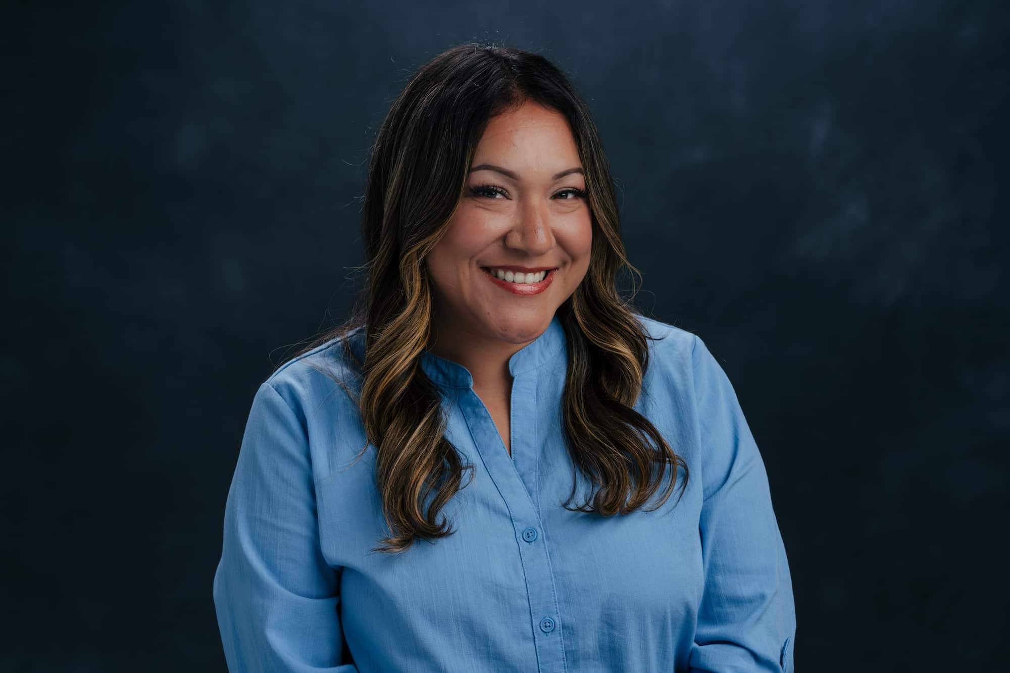 Smiling woman with long dark hair in a light blue blouse against a dark studio backdrop, portrait.