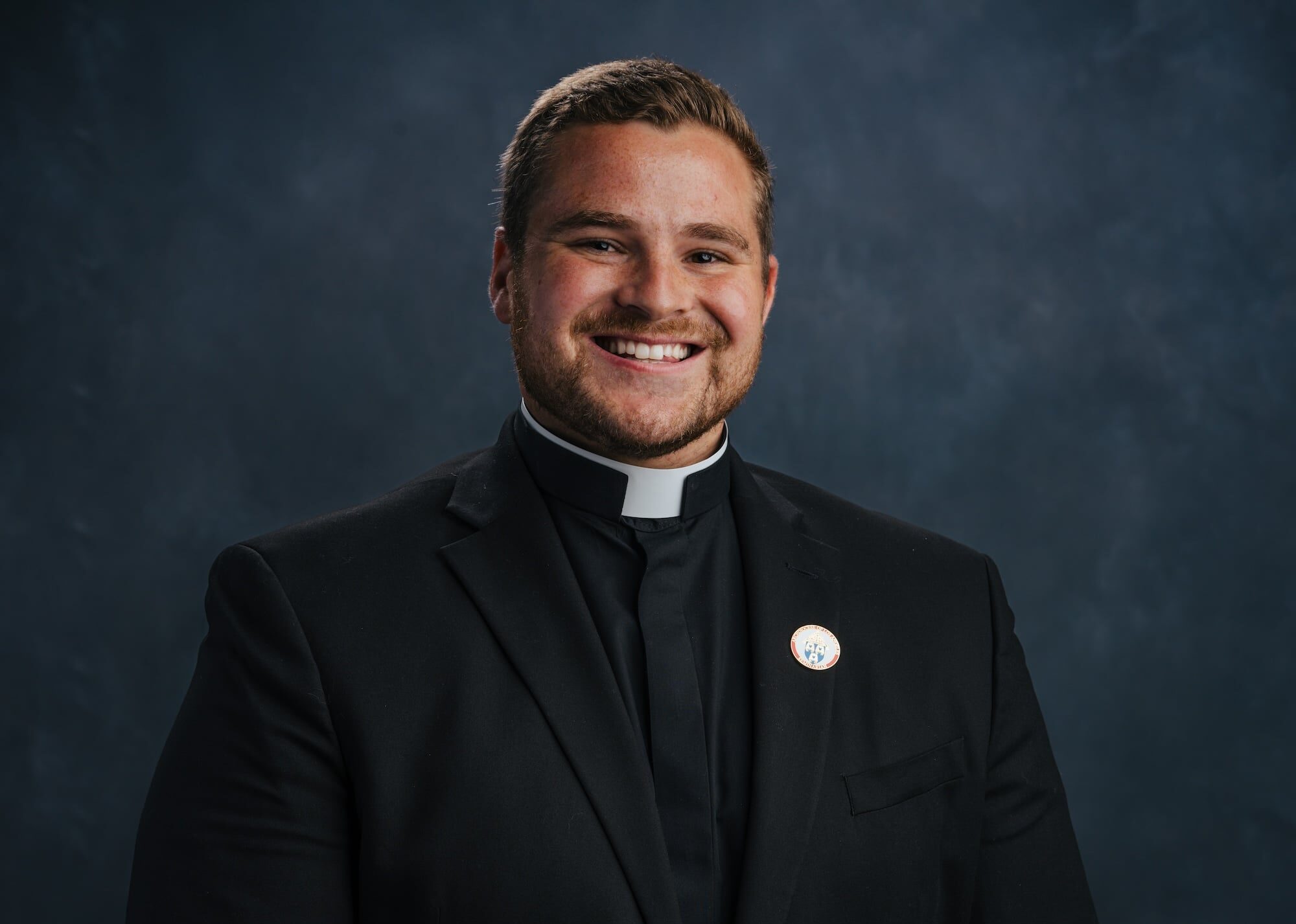 Smiling man in a clerical collar and black suit against a dark textured backdrop