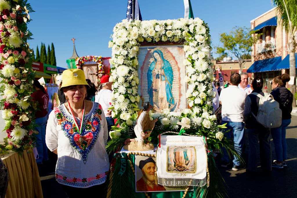 A woman in a traditional embroidered blouse and yellow hat stands beside a large flower-framed religious icon at an outdoor festival.
