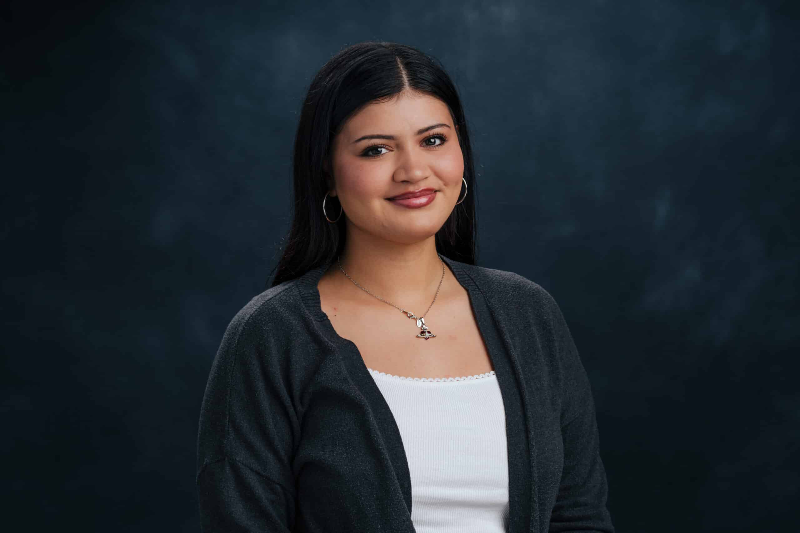 Portrait of a smiling woman with long dark hair, wearing hoop earrings and a silver necklace, against a dark blue backdrop.