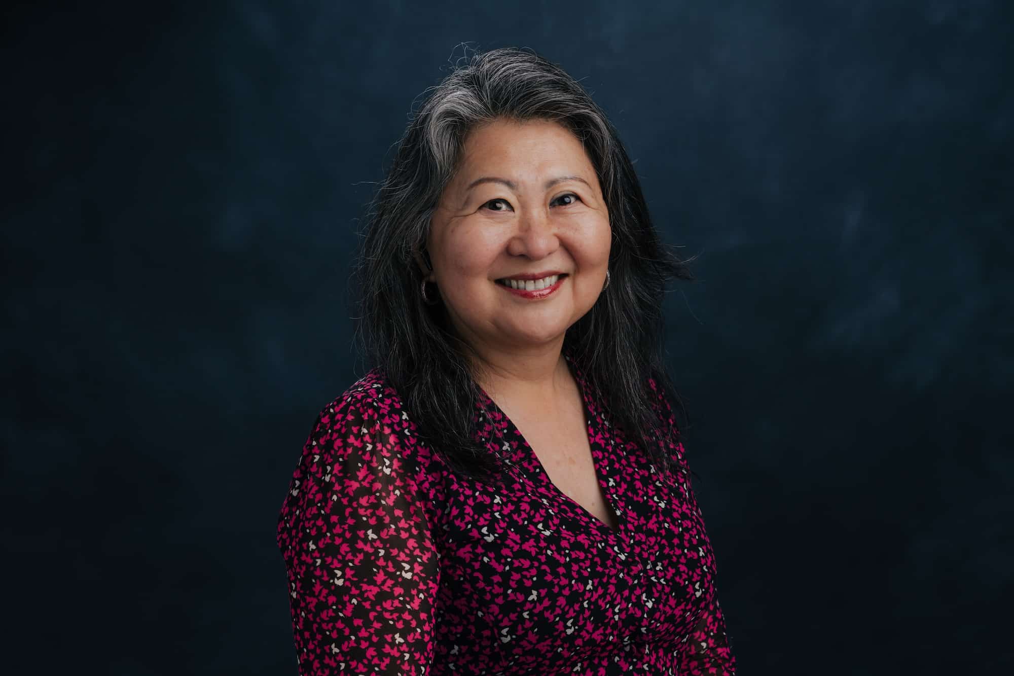 Portrait of a smiling Asian woman with gray hair and a pink and black patterned blouse against a dark backdrop and studio lighting on her face.