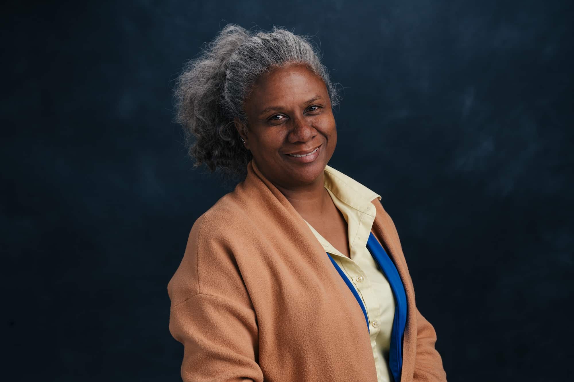 Smiling older Black woman with gray hair, wearing a tan coat over a yellow shirt and blue scarf, against a dark blue backdrop.