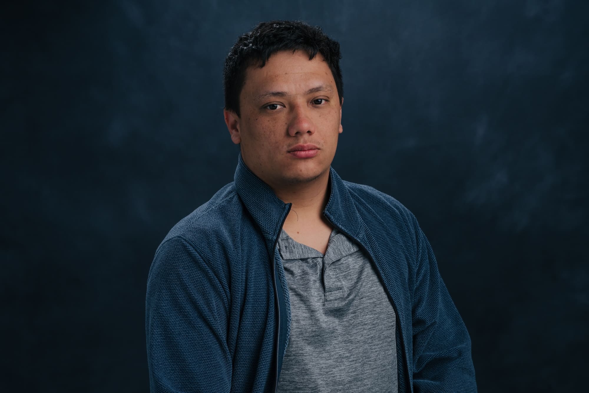Portrait of a young man with short black hair, wearing a gray shirt under a blue textured jacket, against a dark blue studio backdrop.