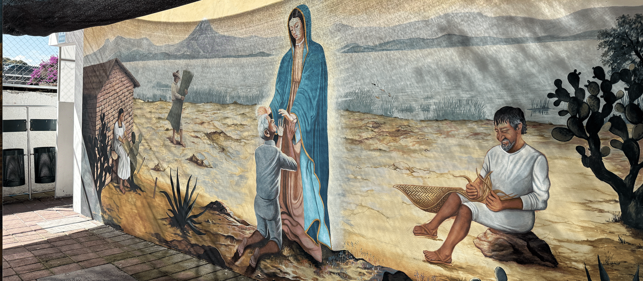 Mural of the Virgin Mary in blue blessing a kneeling man in a desert landscape, with workers nearby.