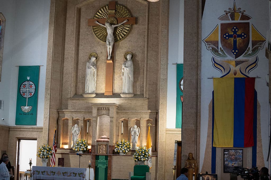 Church interior altar with a central crucifix flanked by two statues, surrounded by floral arrangements and lit candles.