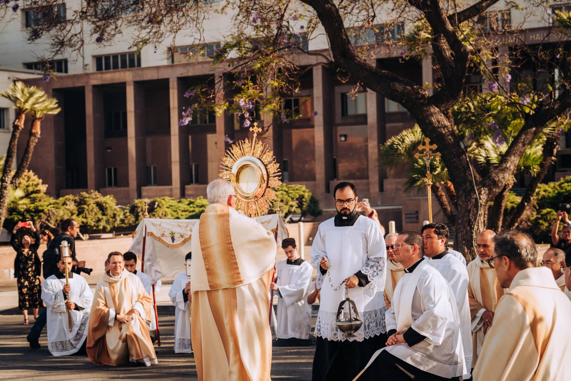 Religious procession outdoors with priests in white robes around a golden monstrance centerpiece.