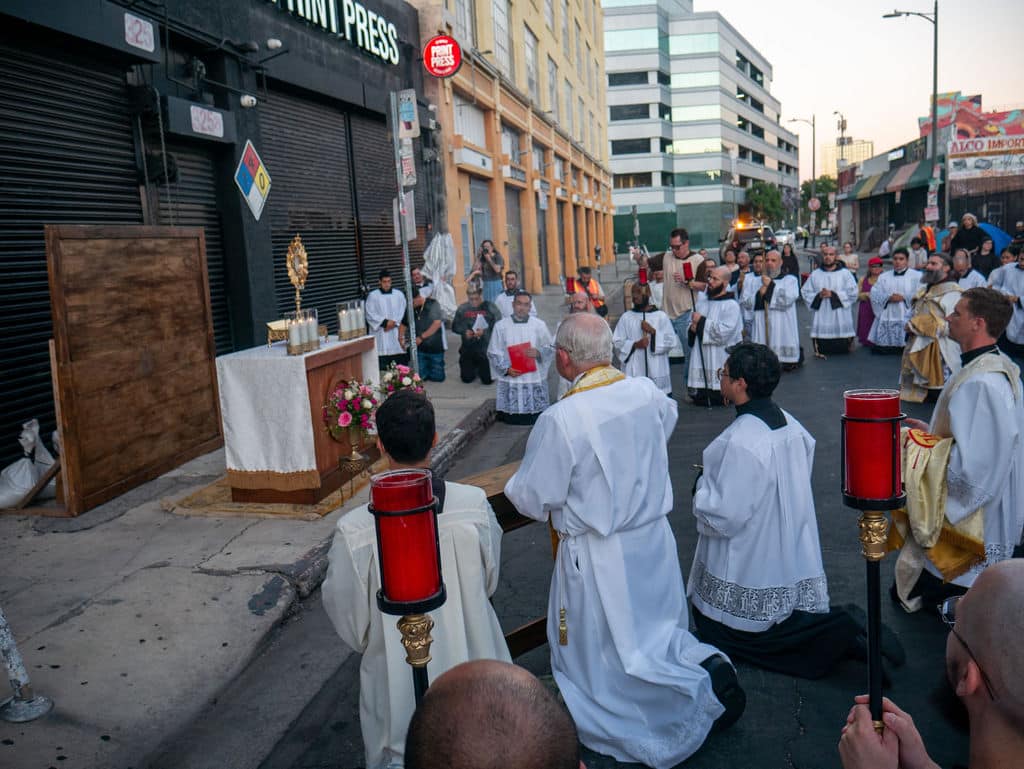 Religious street procession with priests in white robes kneeling before an outdoor altar on a city street, surrounded by onlookers and clergy