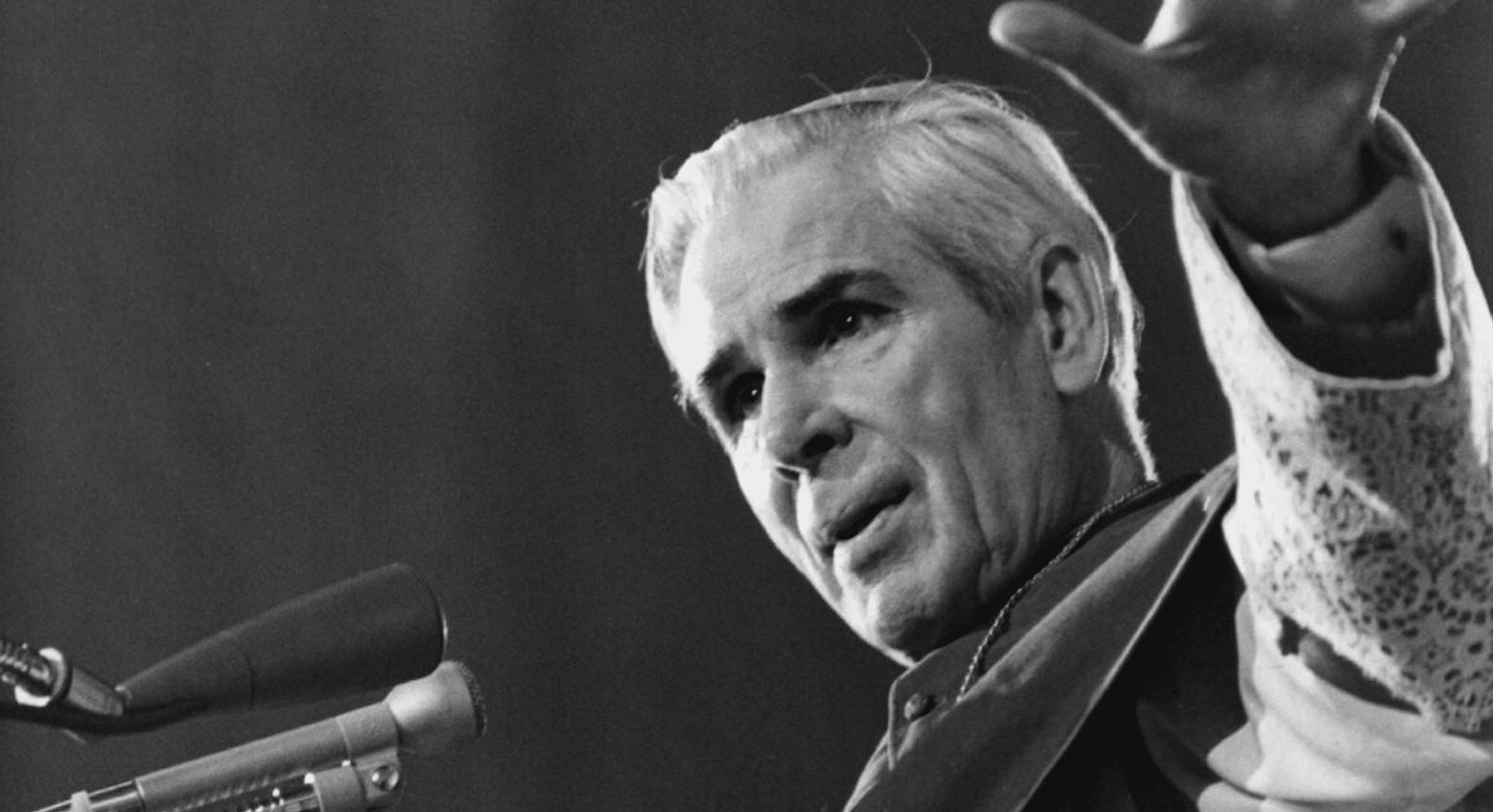 Black-and-white photo of an older man with light hair speaking into a microphone on stage, raising a hand gesture.