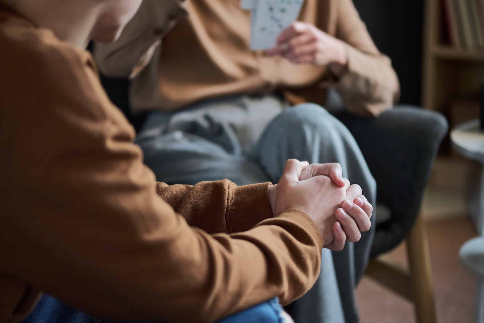 Close-up of two people embracing, focus on intertwined hands in a brown-sleeved arm, indoors in a cozy room (blurry background).